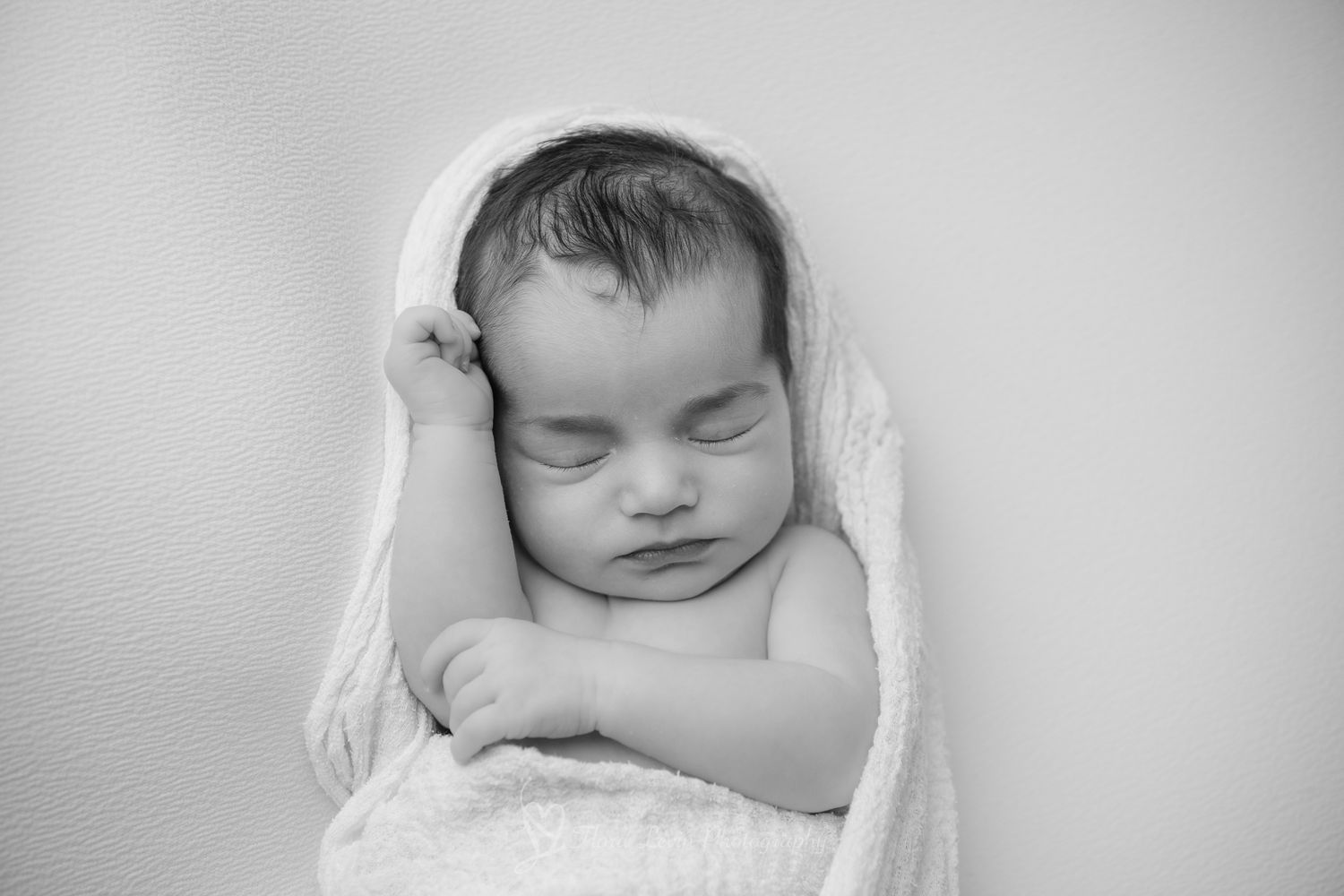 A sleeping newborn baby wrapped in soft white fabric rests peacefully in black and white photography in the Studio of Flora Levin.