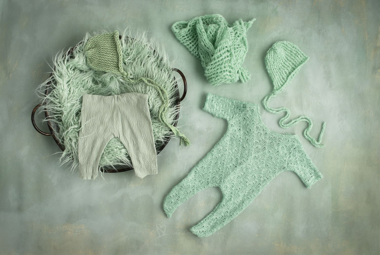 Mint green knitted newborn outfit set with romper, bonnet, booties, and pants displayed on a textured background.