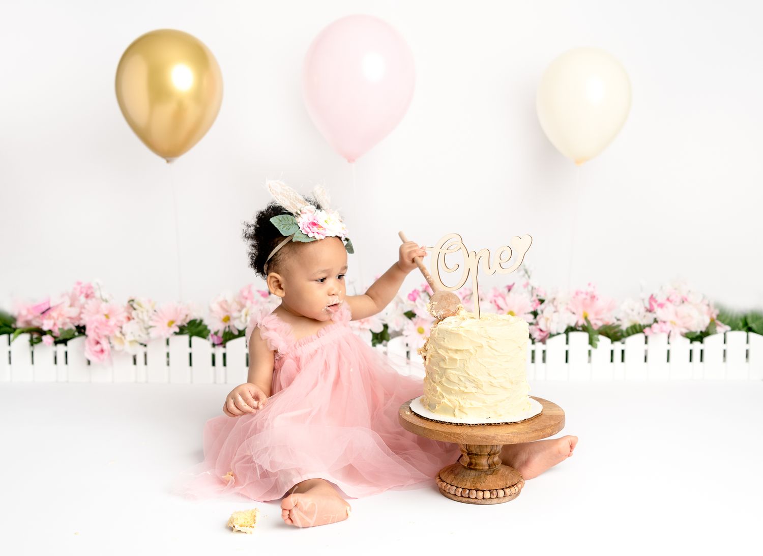 cake smash  session with balloons and garden  , baby girl dressed in pink tulle dress touching the cake with a wooden spoon.