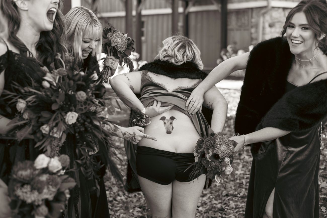 Black and white photo of bridesmaids laughing together while showing off a bridesmaid's lower back tattoo during a wedding.