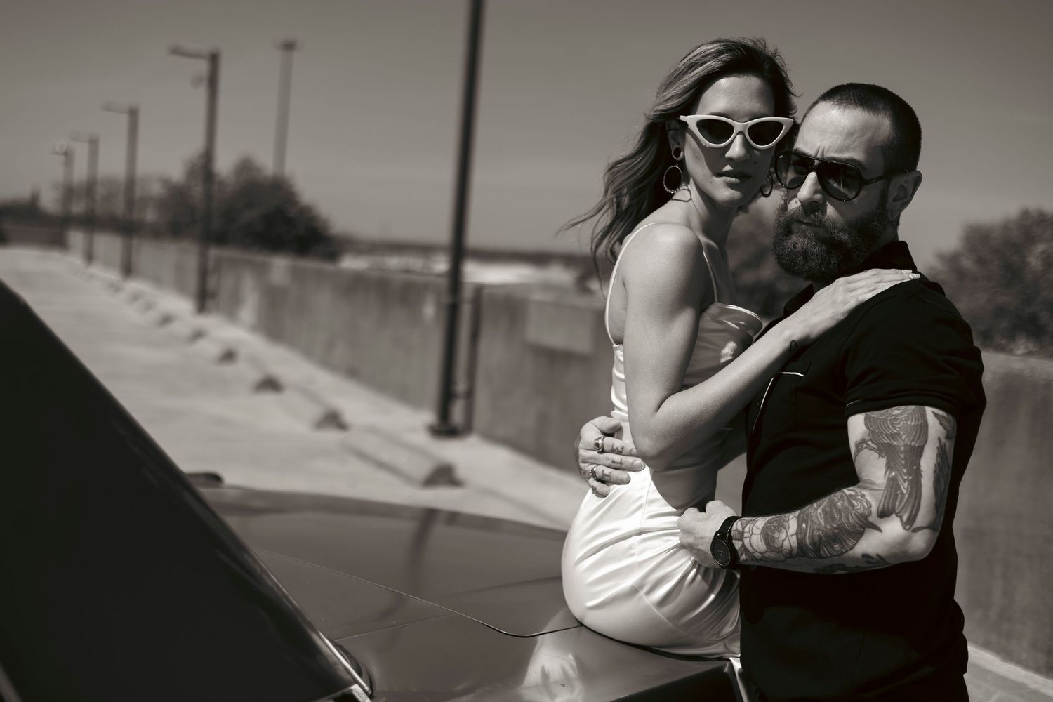 Engagement session photo in black and white of a woman in a white dress and cat eye sunglasses on the hood of a vintage car with her fiancé.