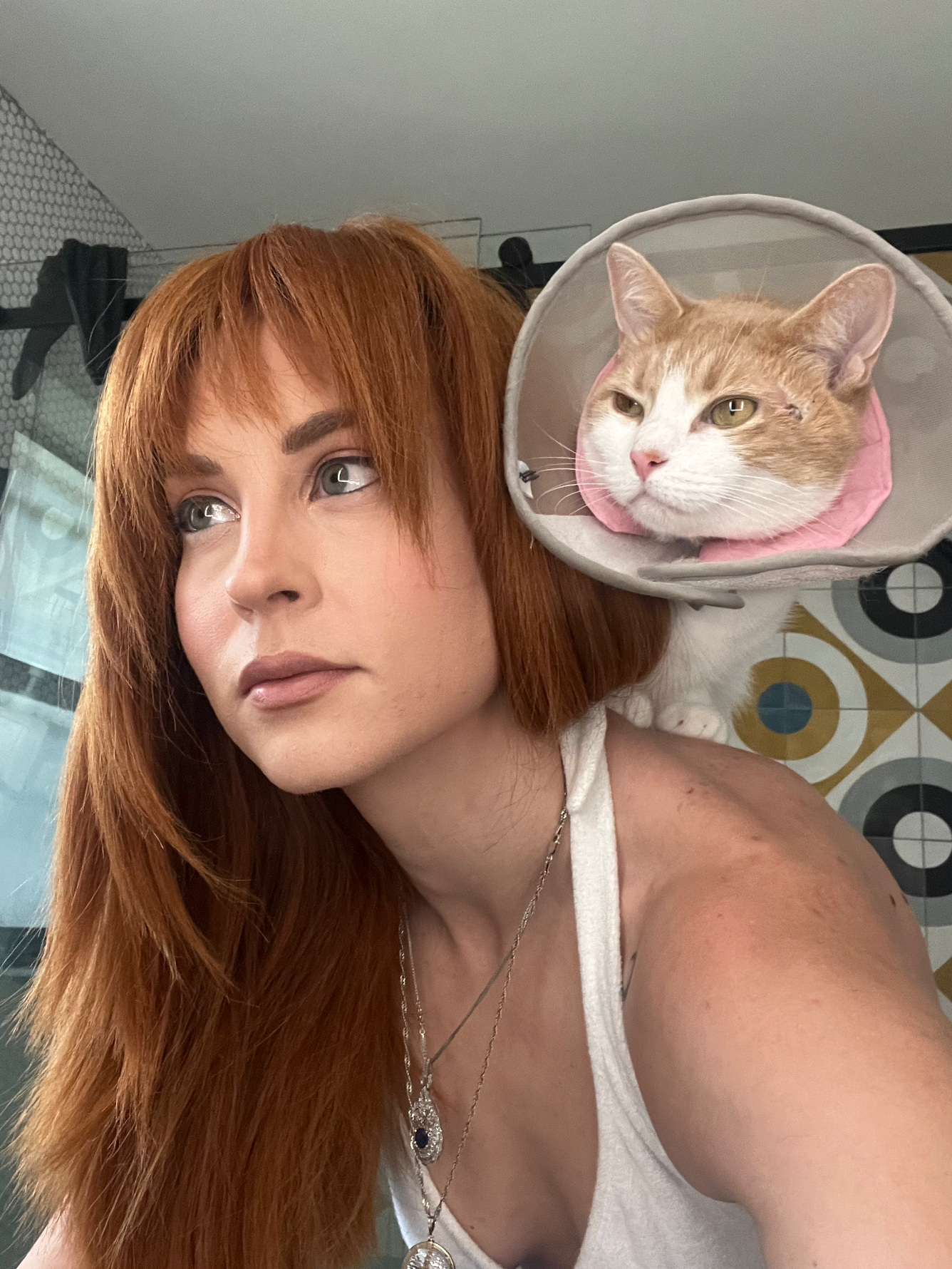 A cat wearing a protective cone collar poses with someone in a white top with red hair.