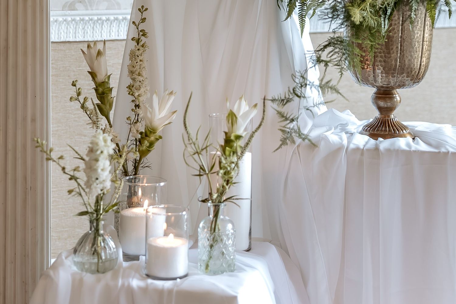 White floral centerpiece arrangements with twigs in glass vases line a rustic wooden table with a lace runner.