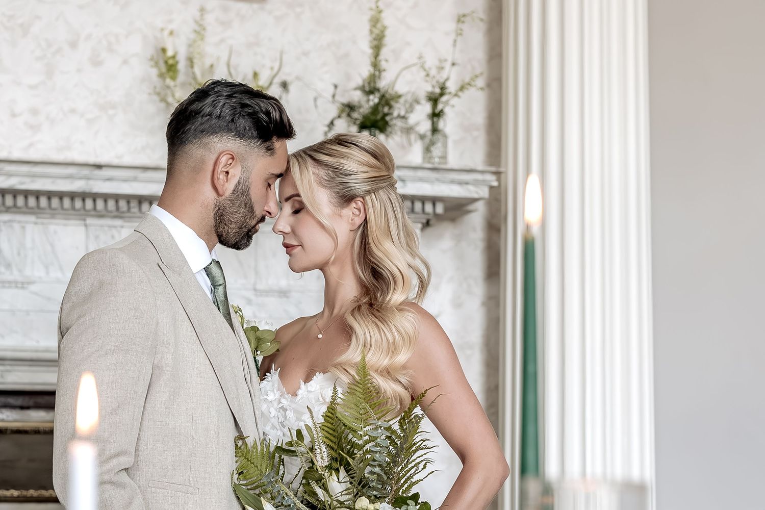 Two people in wedding attire share a tender moment by a fireplace with white and green floral decor and candles.
