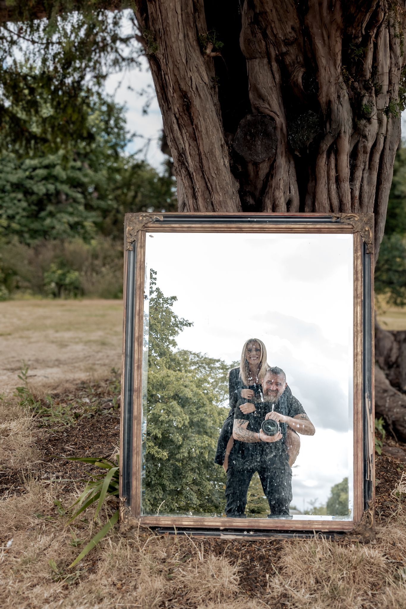 Husband and wife wedding photography team posing with large vintage mirror in outdoor woodland setting.