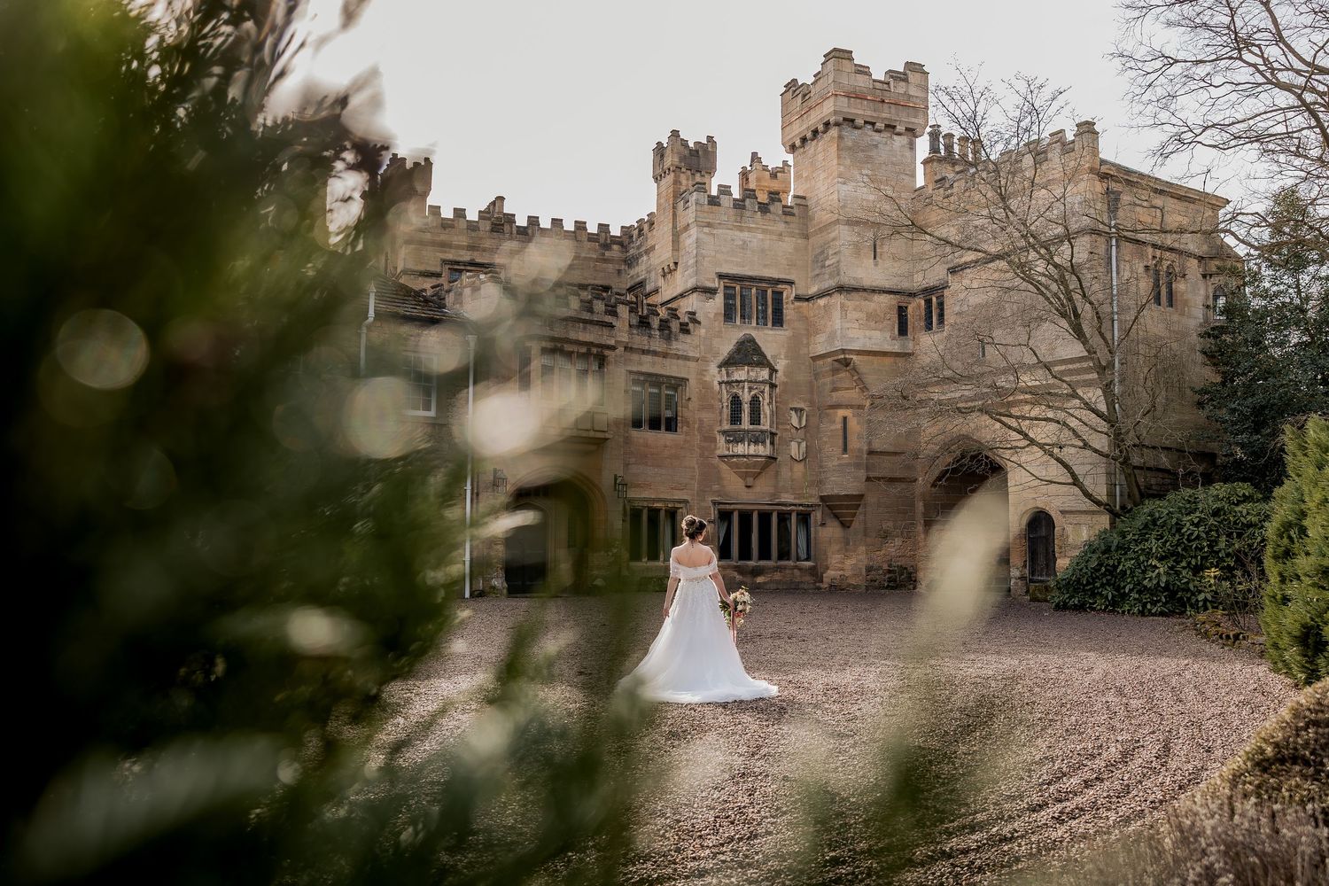 Historic stone castle with Gothic architecture and turrets surrounded by trees with bride posing in courtyard.