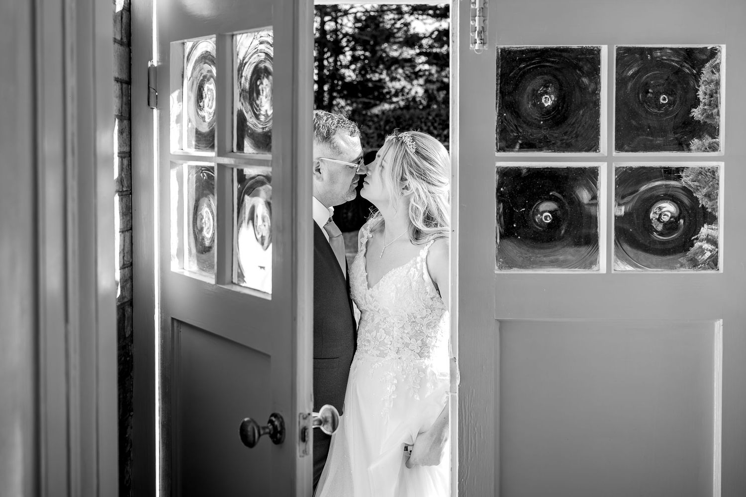Black and white wedding photograph showing a bride in an elegant white dress posing near a window in a vintage setting.