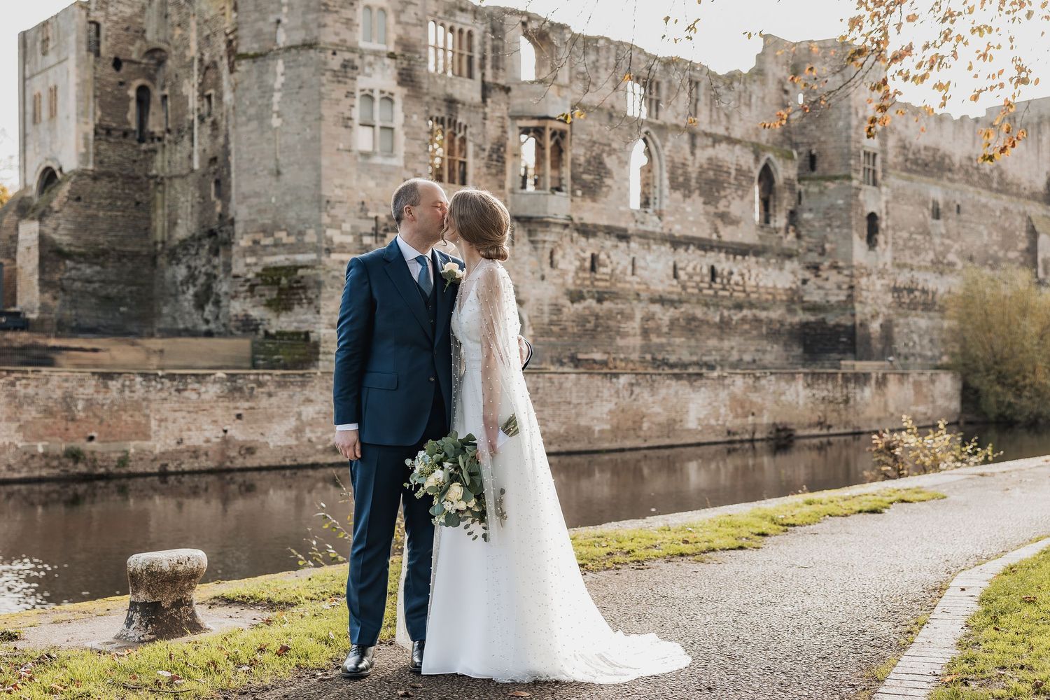 Couple in wedding attire walking along historic stone castle walls on a sunny day.