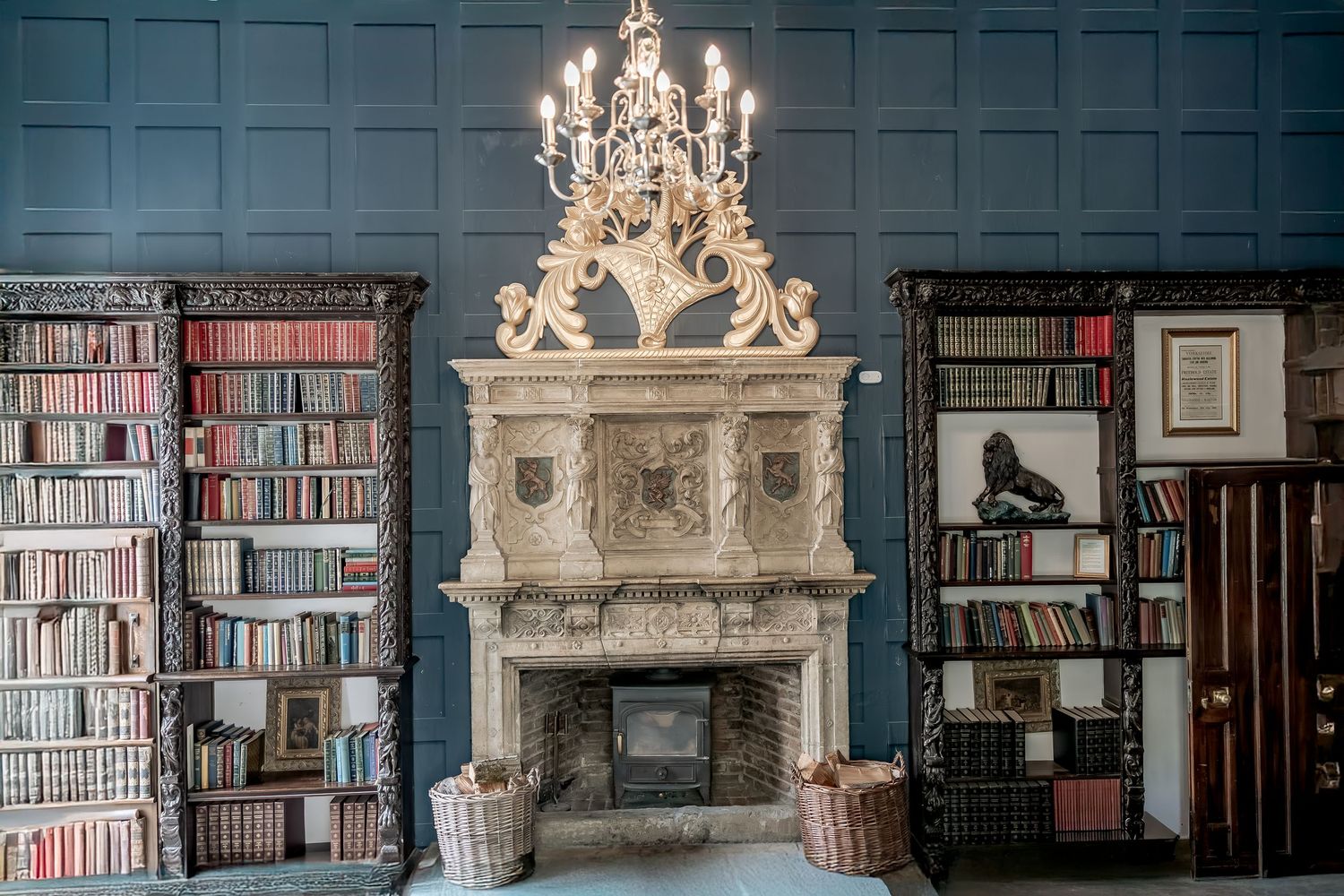 Elegant ornate stone fireplace with crystal chandelier in a vintage library room with blue walls and floor-to-ceiling bookshelves.