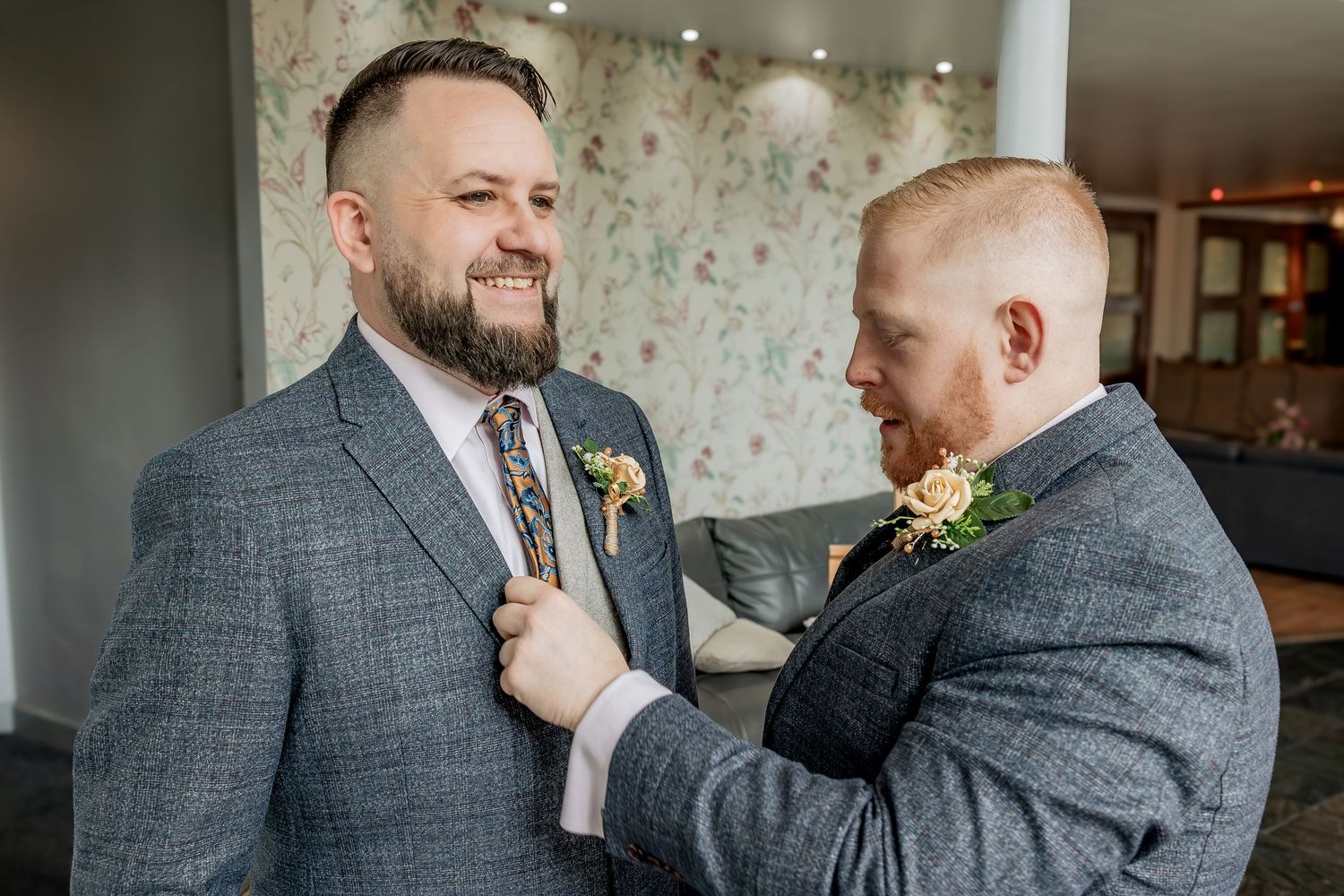 Two people share a joyful moment in formal grey suits while standing indoors with floral wallpaper and decor.