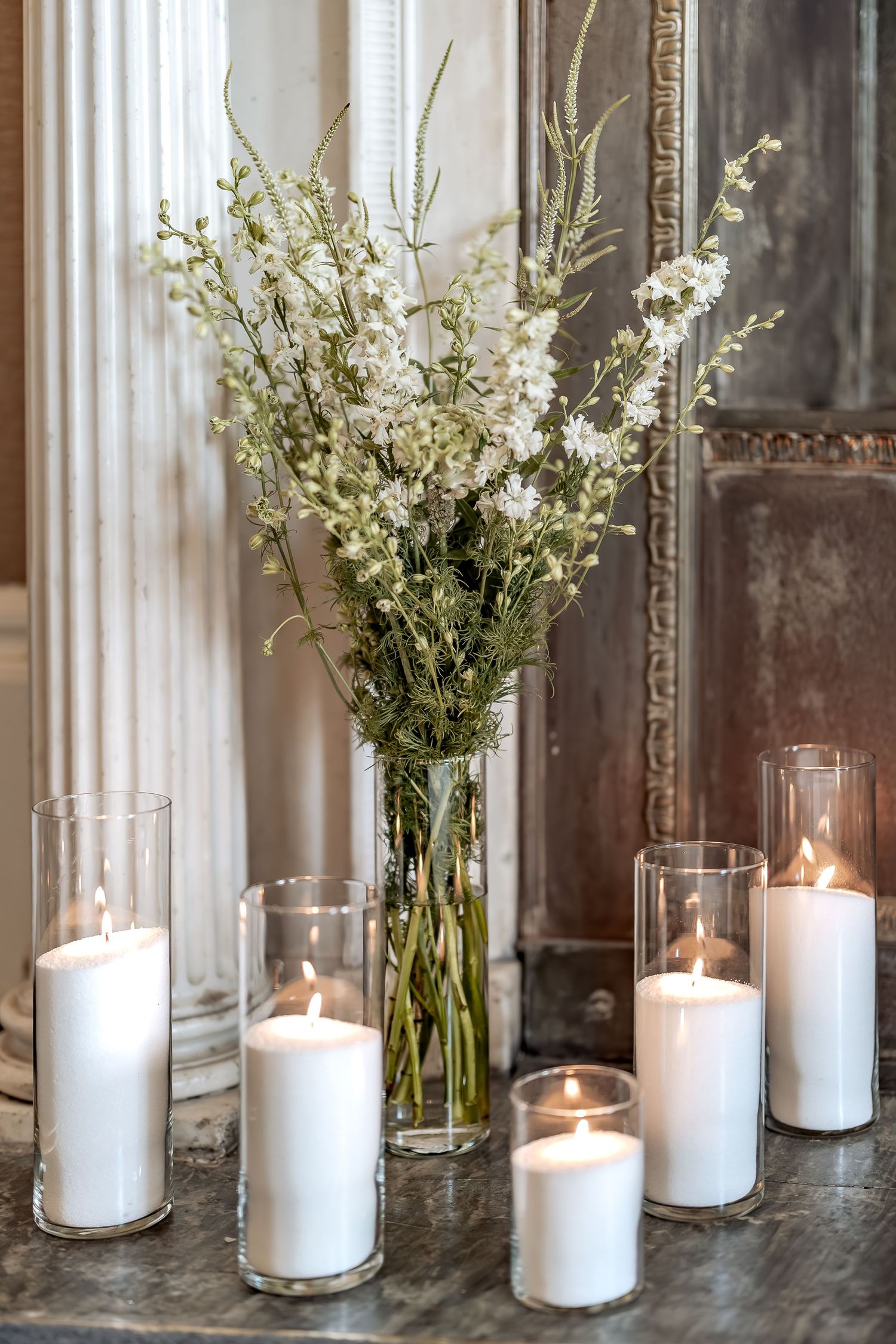 White delphinium flowers in glass vase surrounded by glowing pillar candles in clear holders on gray surface.