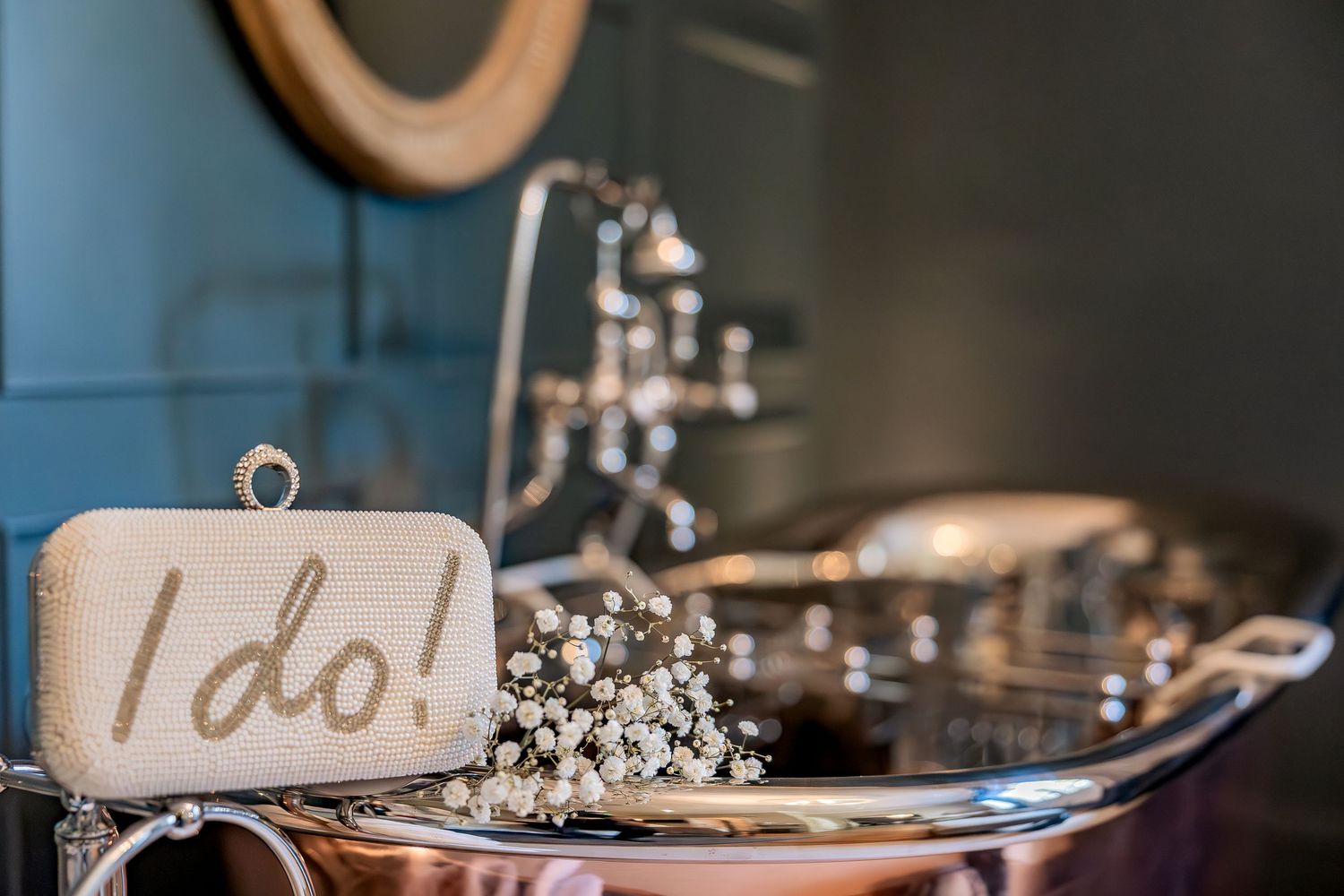 Wedding details with 'I do!' sign, baby's breath flowers, and rings displayed on reflective silver surface in soft lighting.