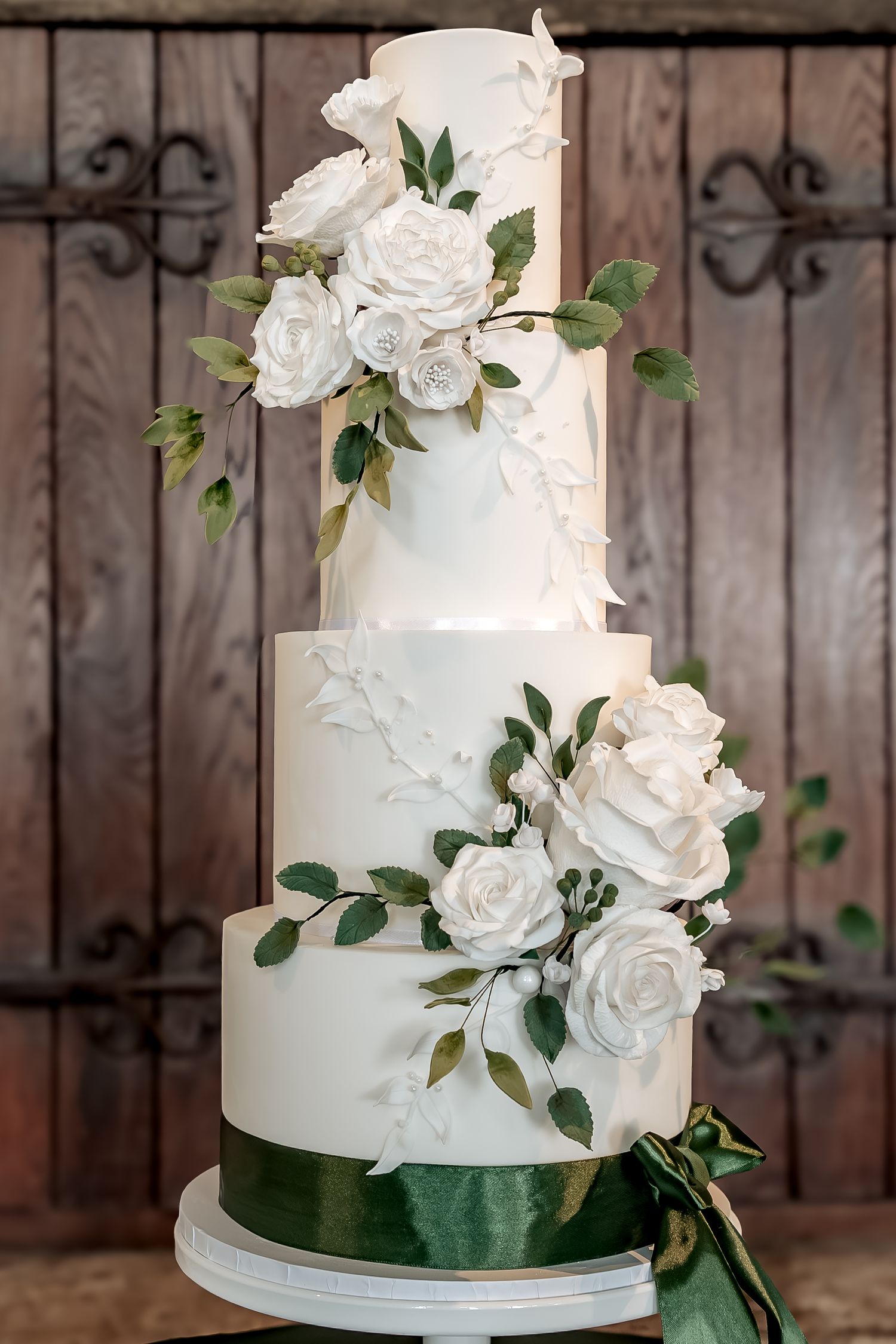 White floral wedding cake with cascading white roses and green foliage against rustic wooden backdrop.