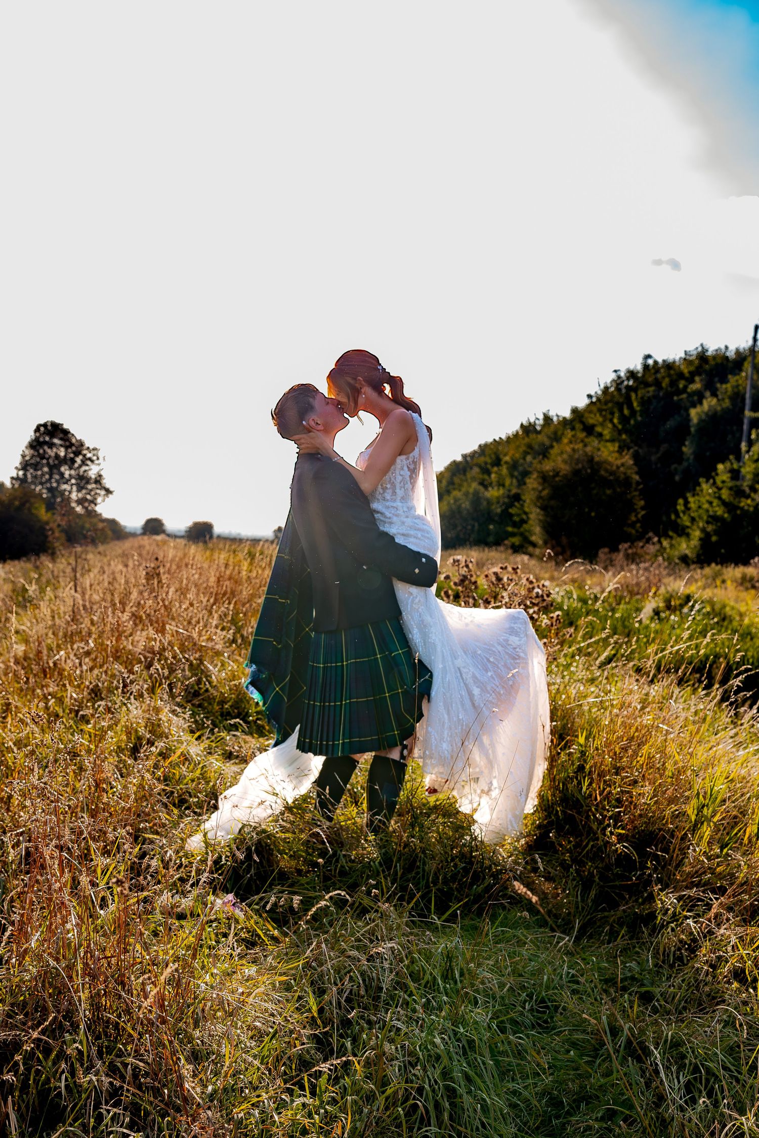 Wedding couple shares an intimate moment at sunset in a grassy field wearing traditional Scottish attire and white gown.