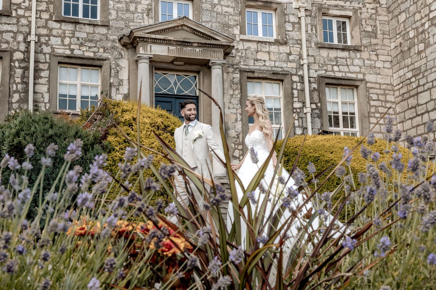 A couple in wedding attire stands in front of a stone mansion with ornate windows and a blooming flower garden.