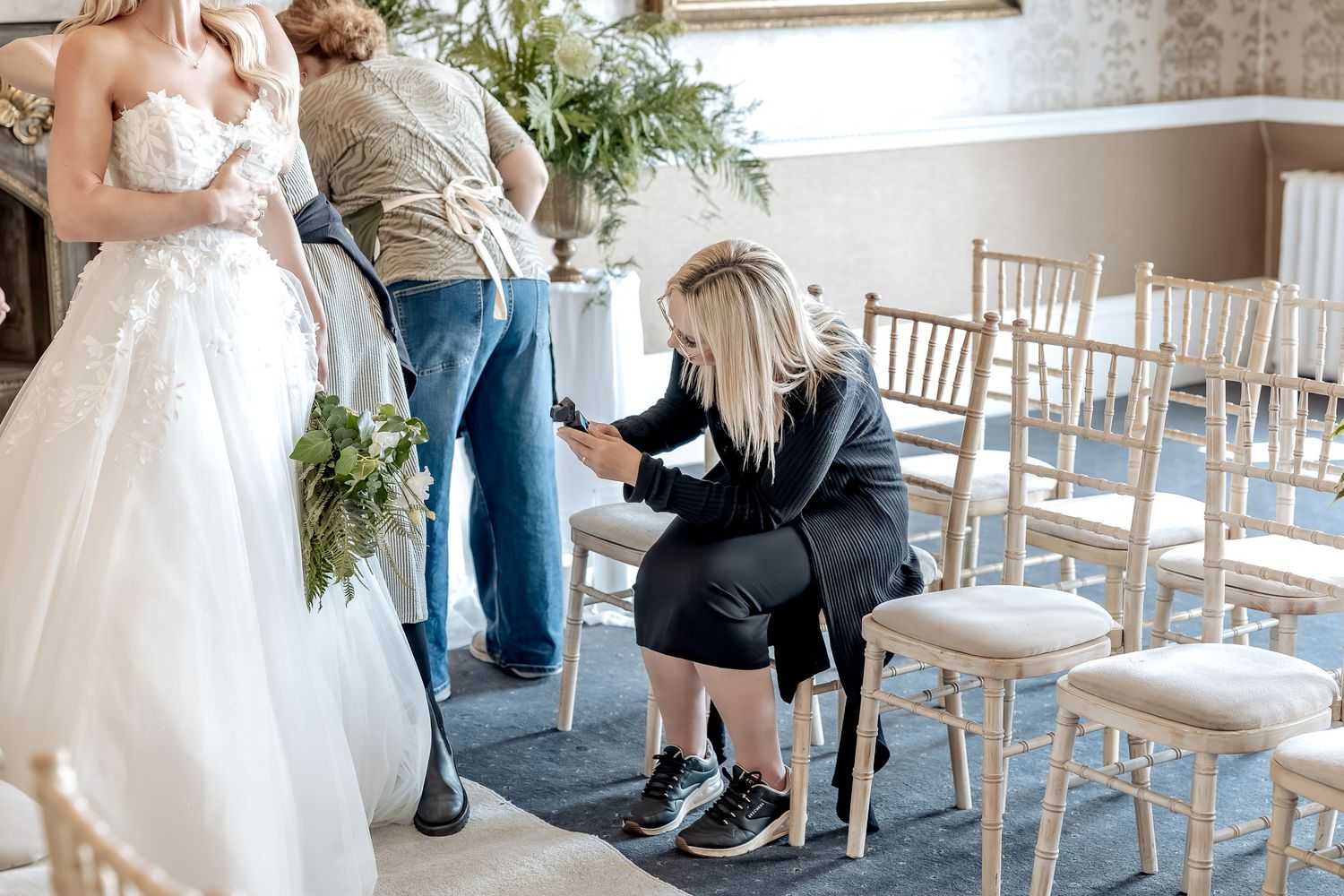 A wedding dress is being adjusted in a room with white chiavari chairs and floral decor, whilst being captured on an osmo pocket 3.