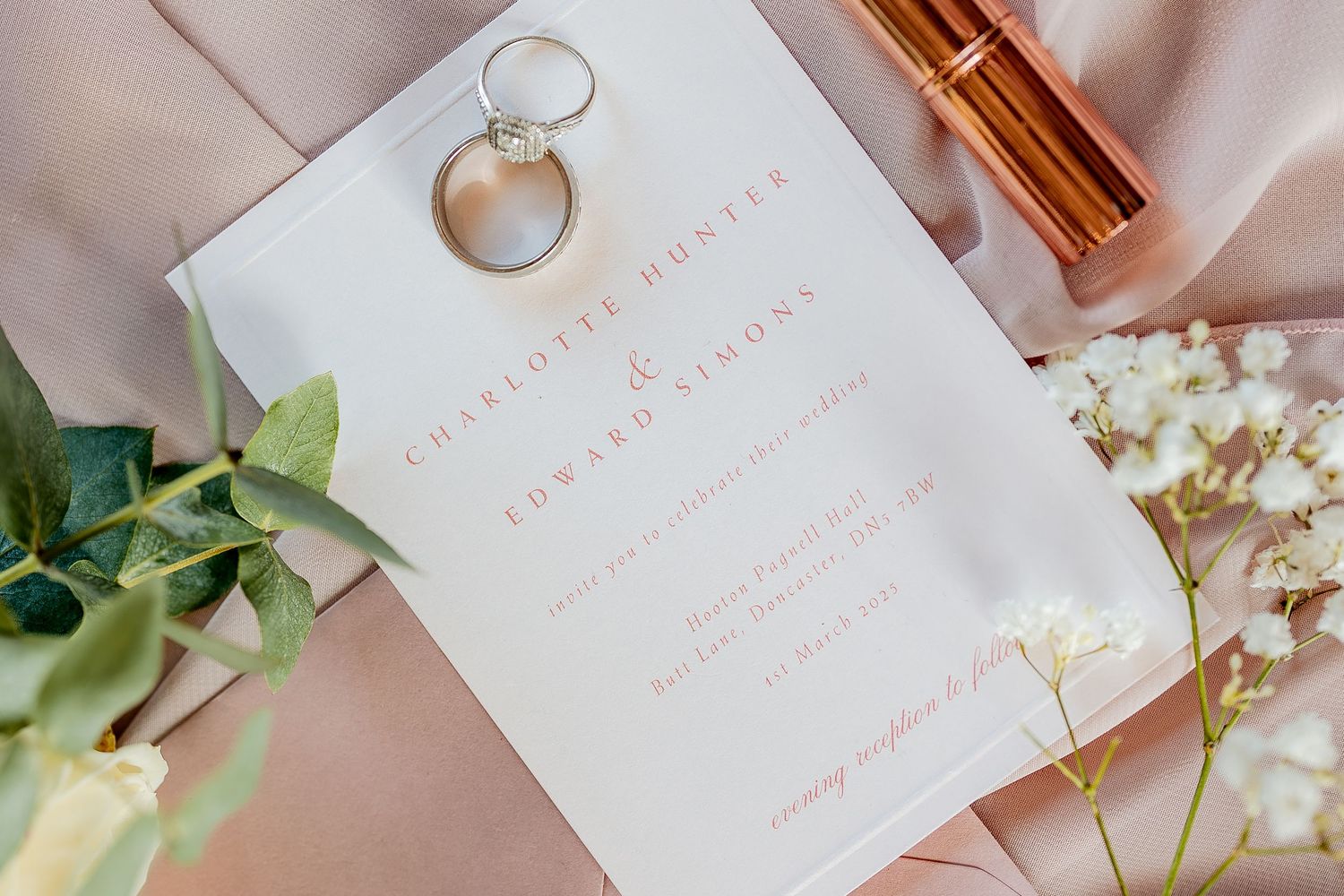 Elegant wedding details flatlay with invitation, gold rings, pink accents, and delicate white flowers on soft fabric.