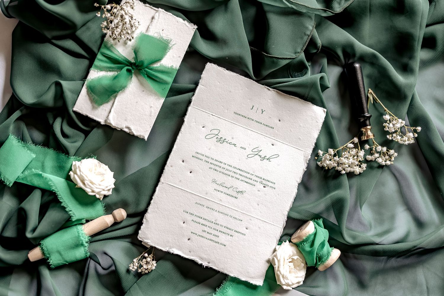 Wedding invitation surrounded by green decorative accents, white floral elements, and wrapped gifts on emerald green silk fabric.