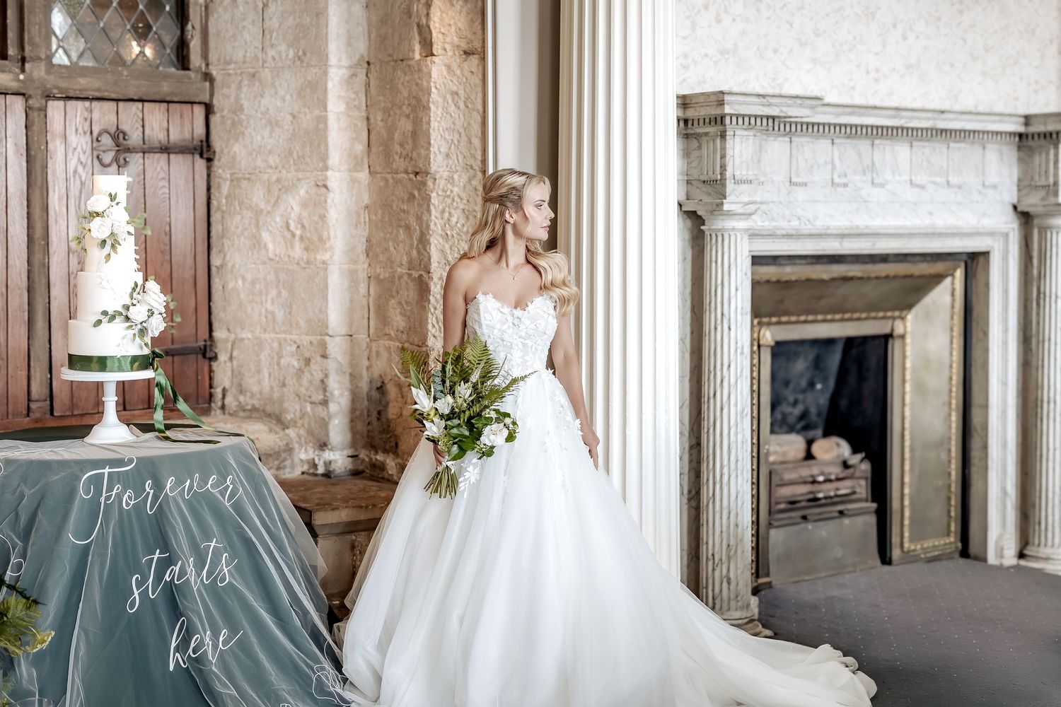 An elegant wedding dress is showcased in a historic room with stone columns and a fireplace.