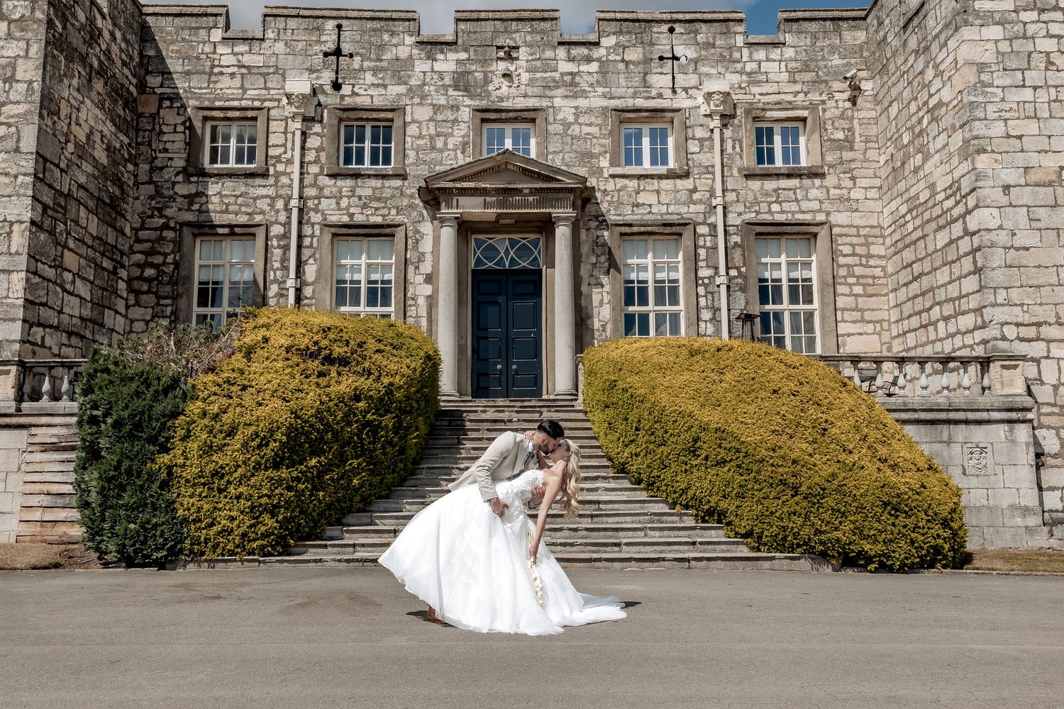 Two figures in wedding attire embrace on grand stone steps leading to a historic building with manicured green bushes.