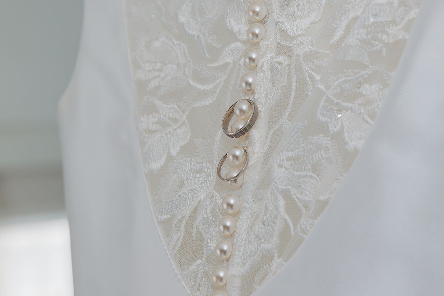 Close-up detail of delicate white lace wedding dress fabric with pearl button embellishments, with wedding rings.