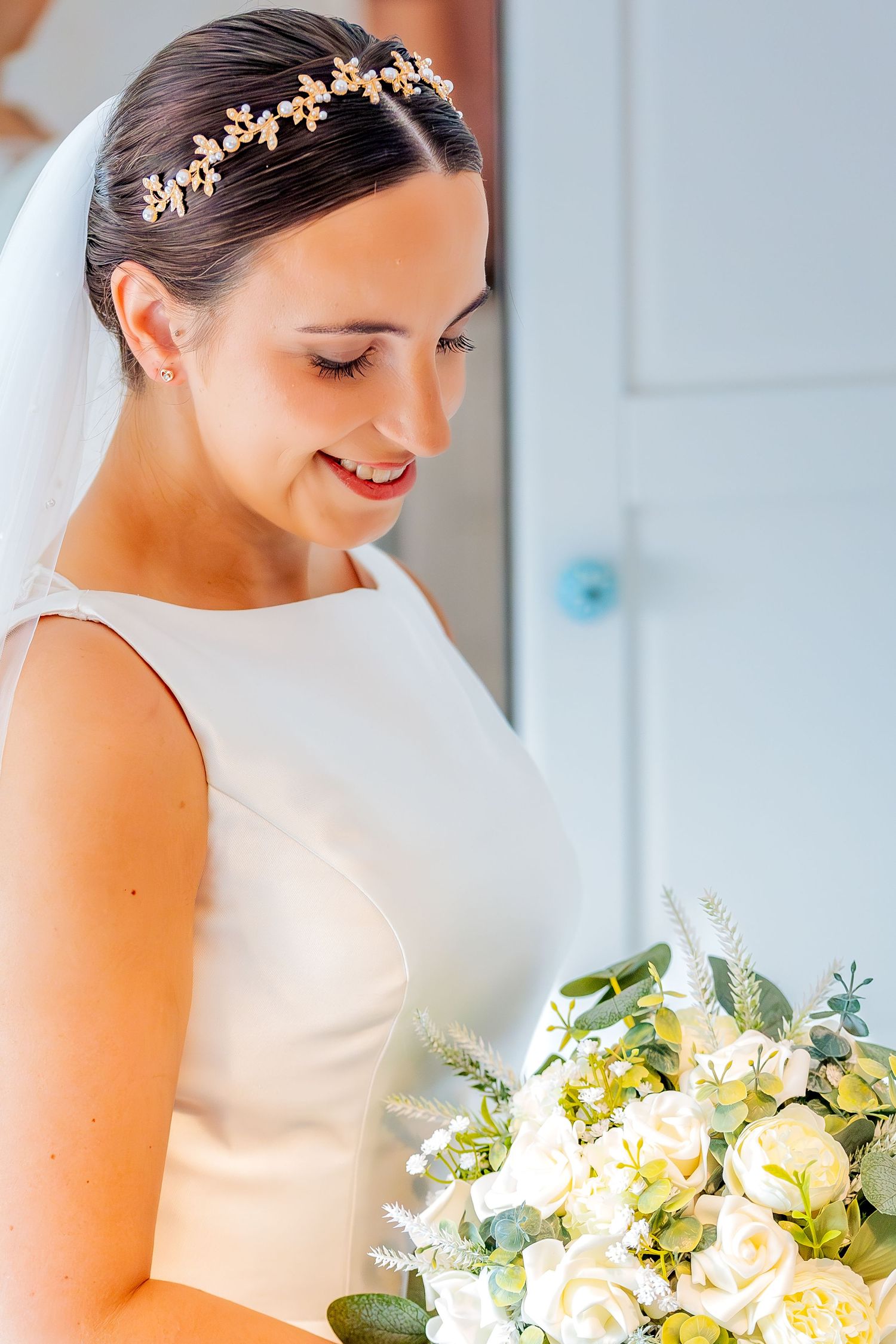 Elegant bride in sleek white dress holds cream and white bouquet while wearing delicate gold leaf hair accessory.