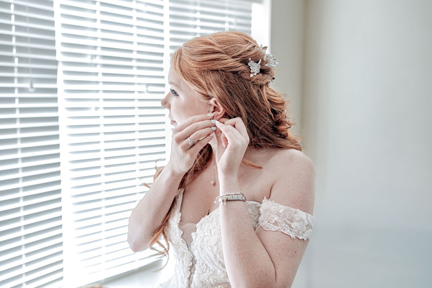 A bride in a white lace wedding dress poses near venetian blinds in soft natural light.
