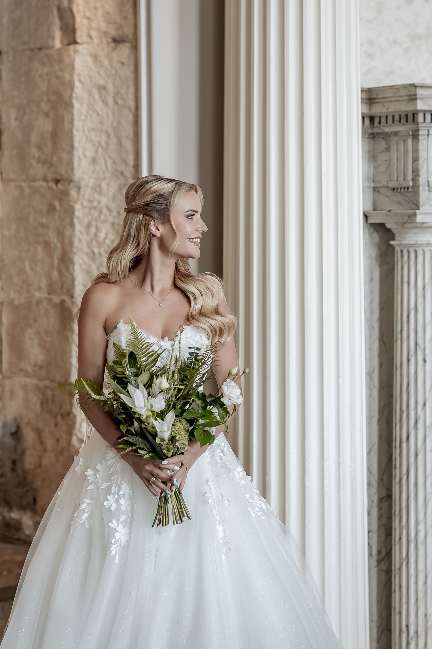 Bride in white strapless wedding gown with floral appliques poses elegantly between classic stone columns.