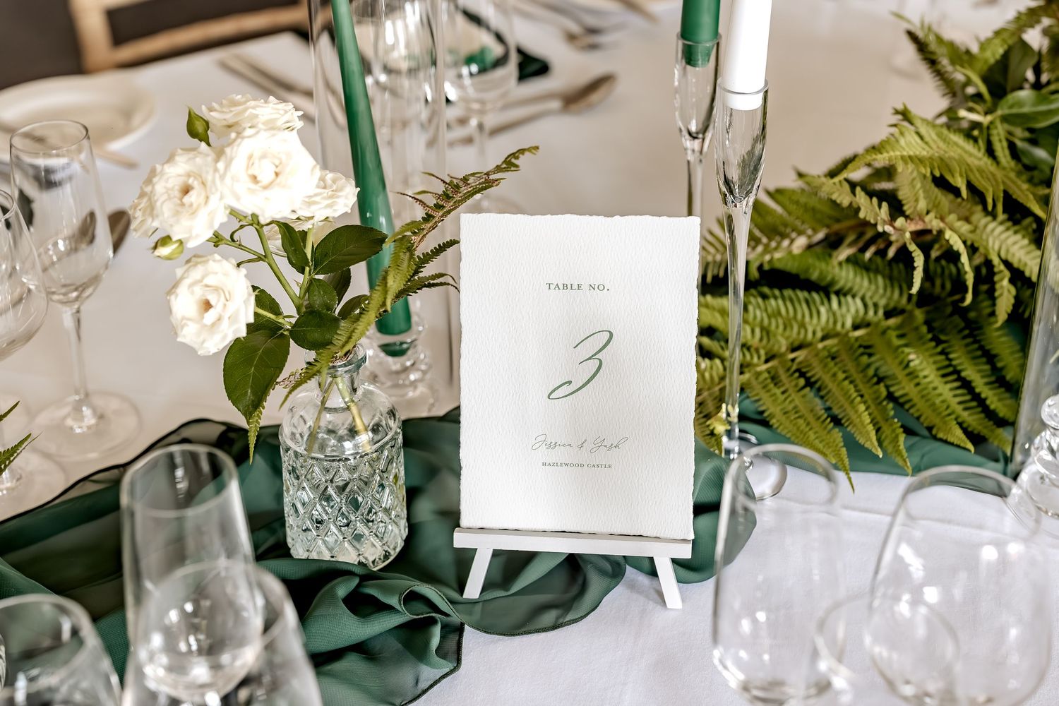 White floral centerpieces with greenery on a table setting decorated with glass vases and candle holders for a wedding reception.