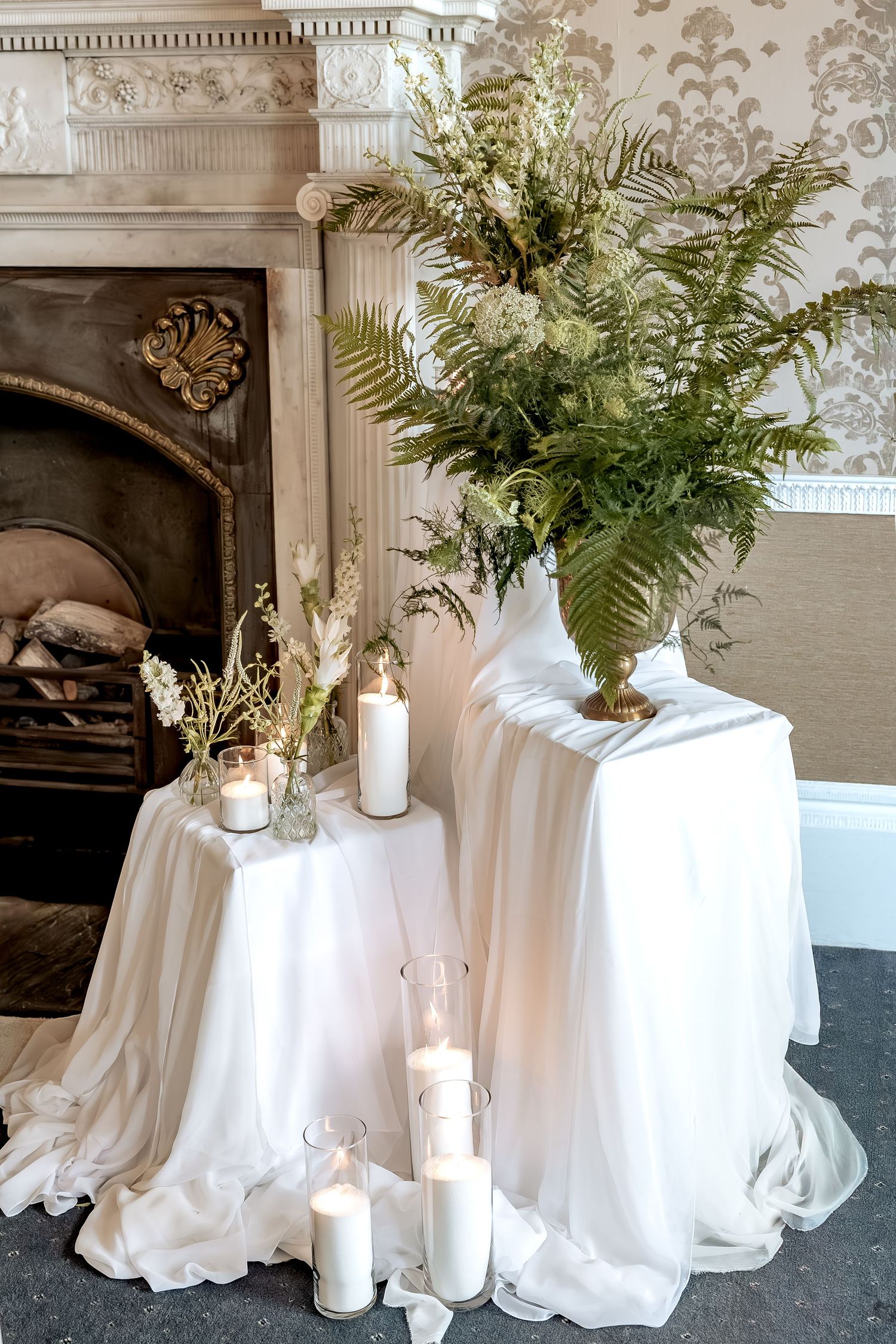 Elegant white draped table with fern arrangement and glowing candles beside an ornate fireplace with decorative wall details.