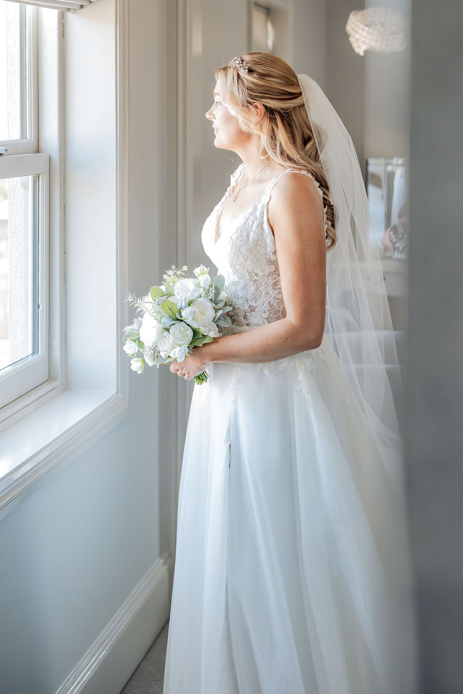 A bride in a flowing white wedding gown stands by a window in soft natural light creating an ethereal bridal portrait.