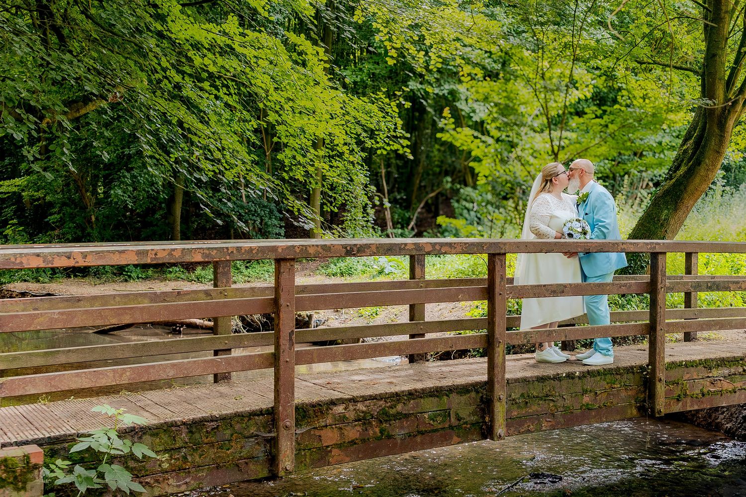 Wooden footbridge stretches across a scenic forest stream surrounded by lush green trees and foliage, with newly weds embracing