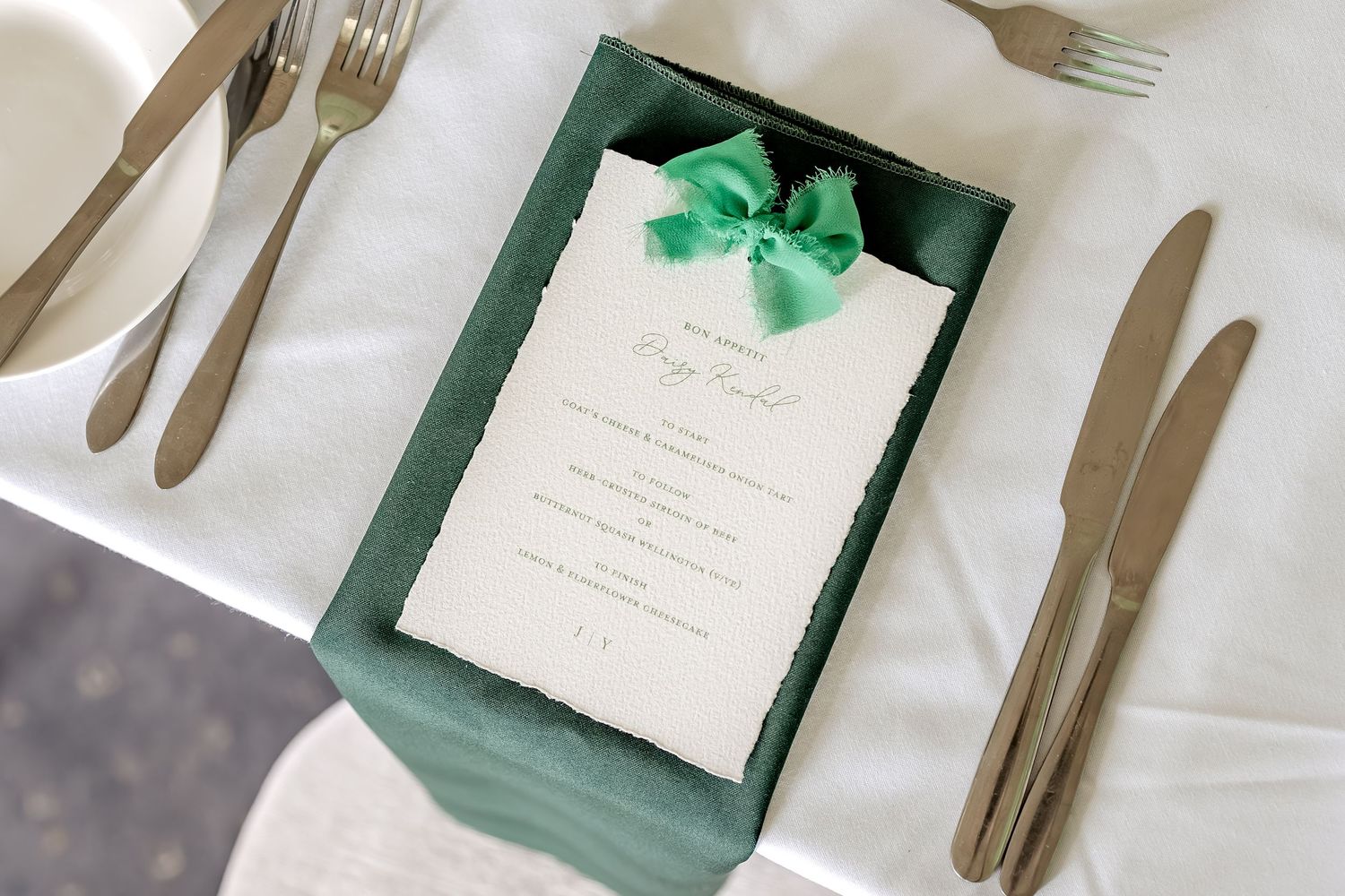 Elegant wedding menu card with emerald green bow displayed on white tablecloth with gold cutlery.