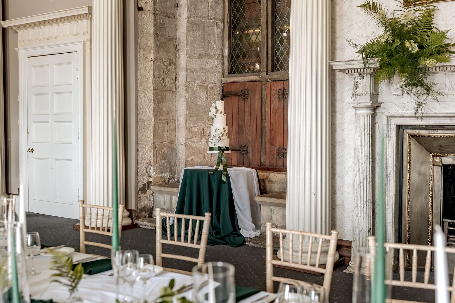 Elegant wedding reception table setup with white chairs and emerald green decor and wedding cake in a historic stone venue.