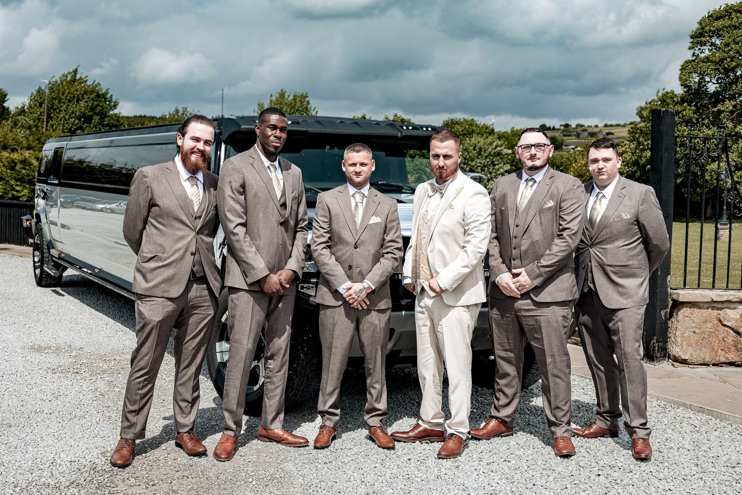 Wedding party stands together in front of a stretch limousine wearing coordinated gray and beige suits.