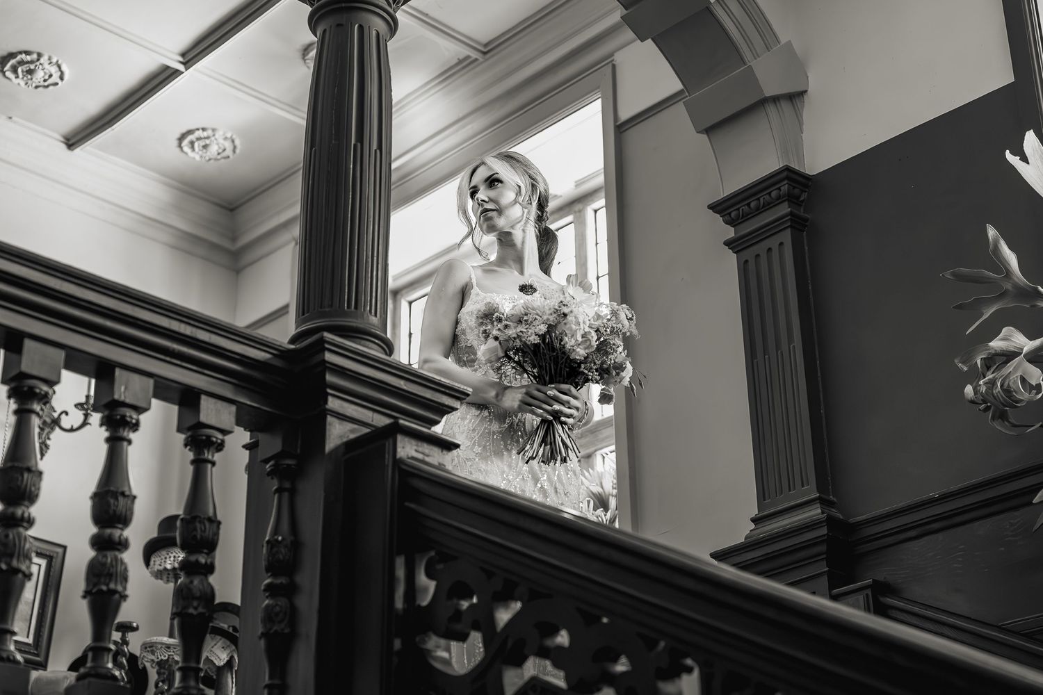 A black and white photo of a bride holding a bouquet while standing at the top of an elegant staircase with dark columns.