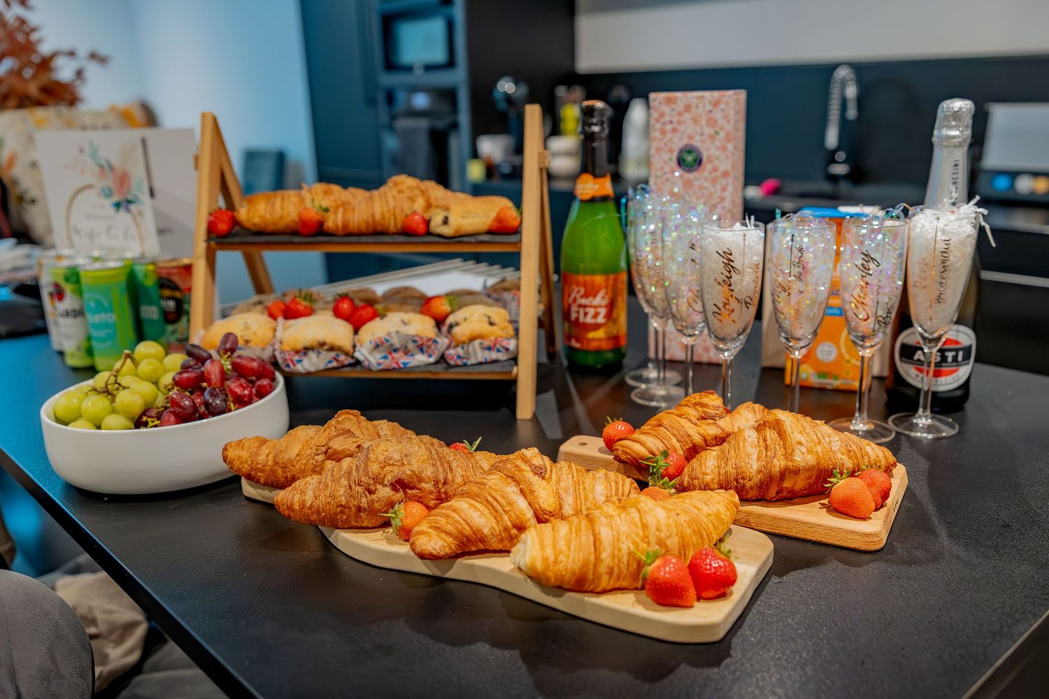 Fresh baked croissants arranged in rows on dark surface with colorful plates and beverages in background.