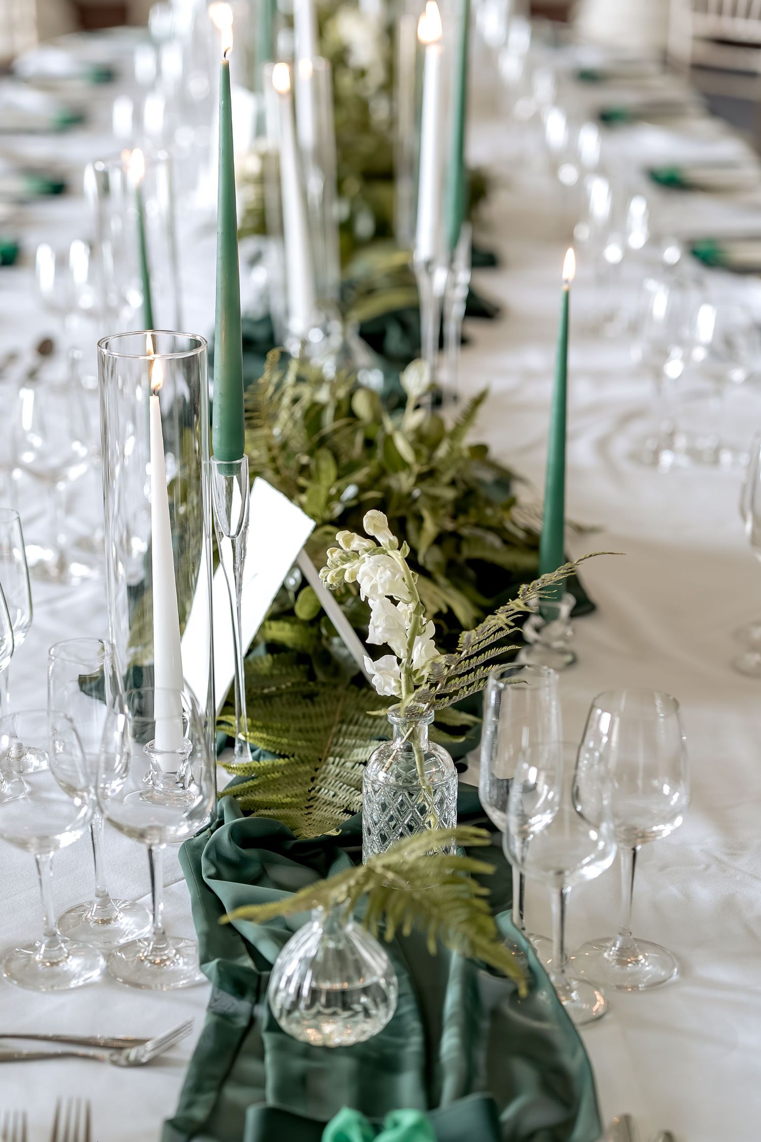 Elegant table setting with tall green candles, wine glasses and greenery runner on white tablecloth for formal event.