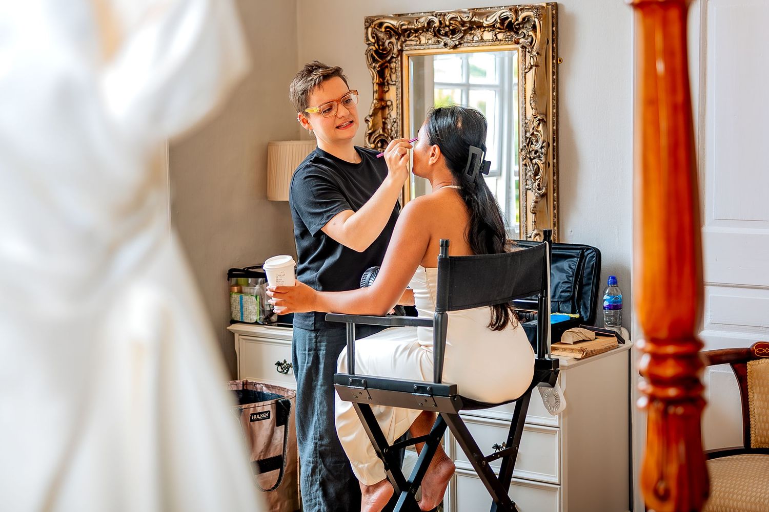 A makeup artist applies cosmetics to a bride seated in a professional chair with beauty tools and supplies nearby.