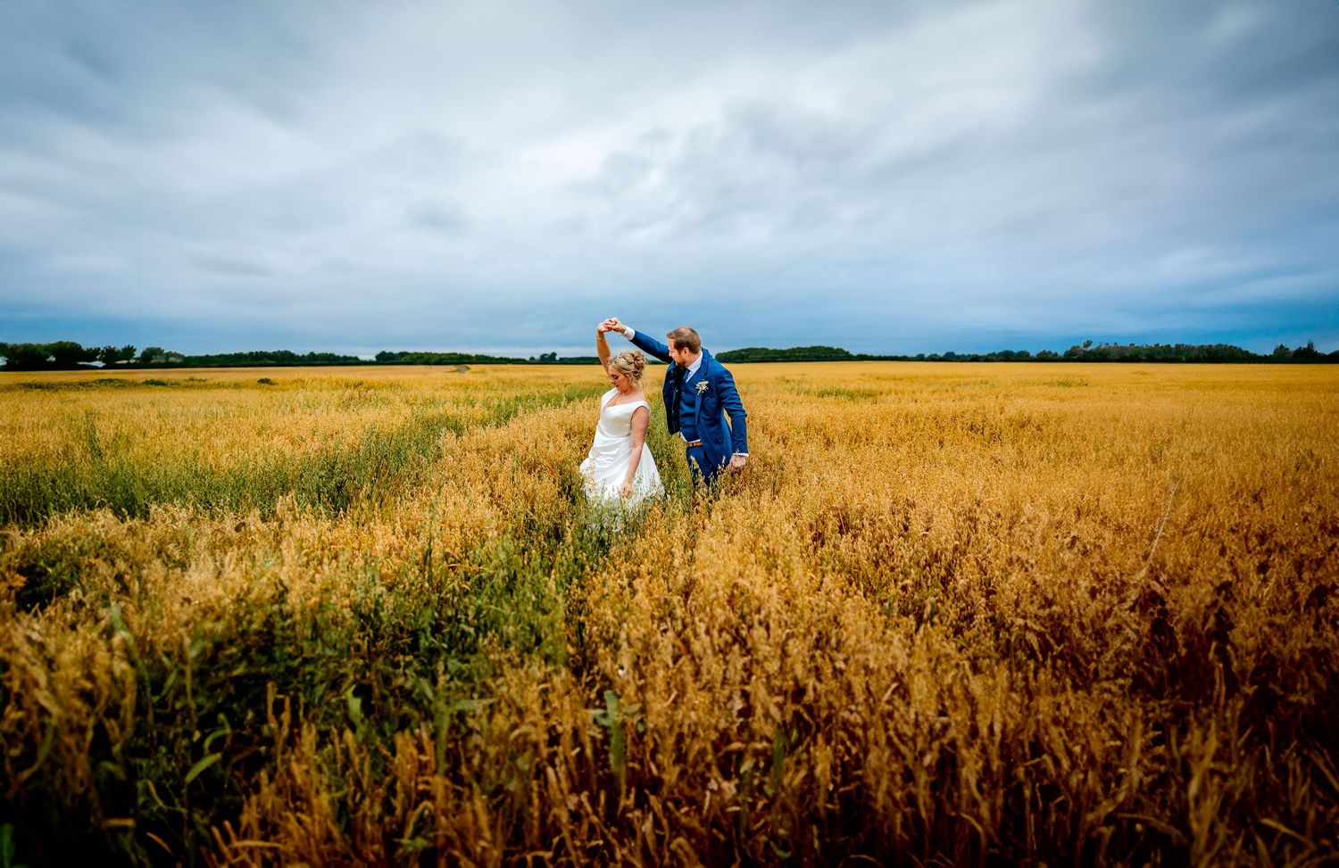 A joyful couple share a dance in golden wheat field under dramatic cloudy skies at sunset.