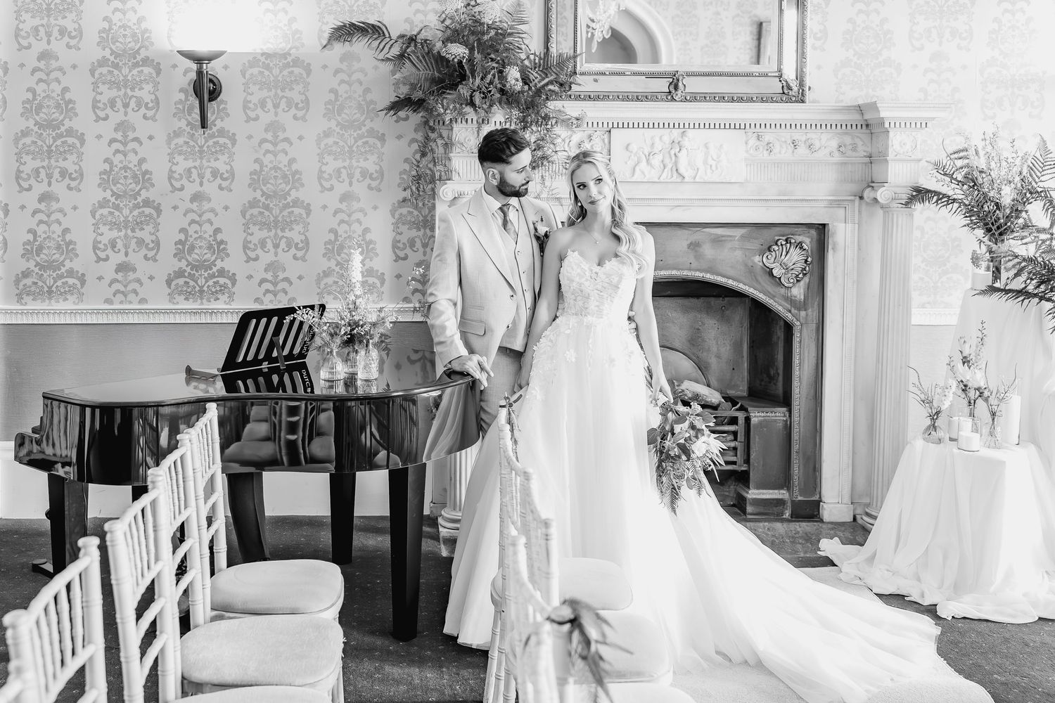 A black and white wedding photo showing elegant couple sharing a moment next to a piano in an ornate venue.