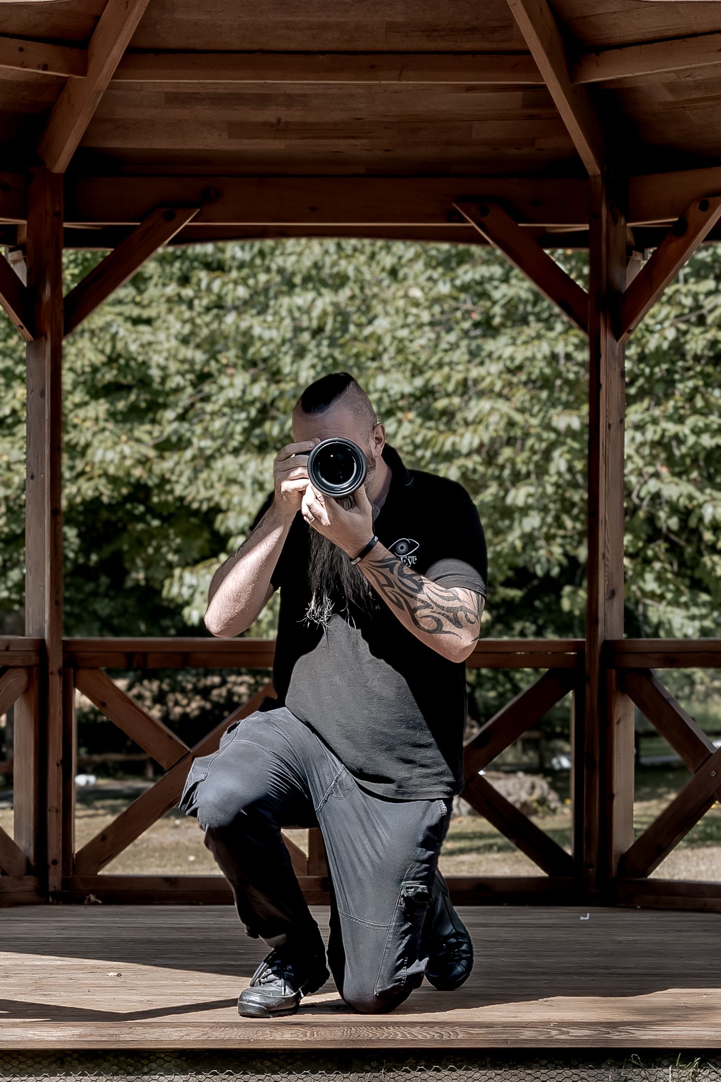 Photographer in casual attire kneeling on wooden deck while taking photo with professional camera in outdoor setting.