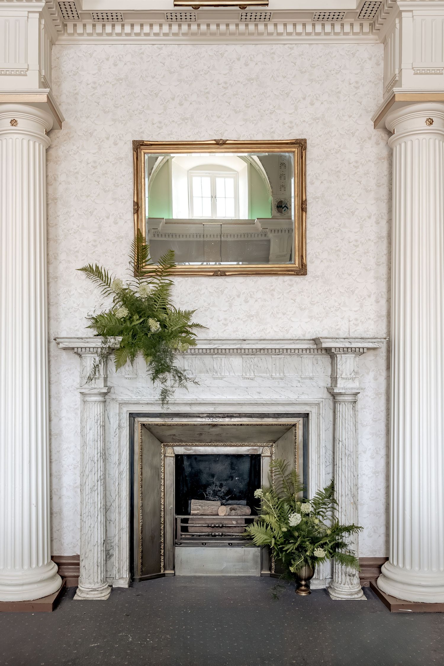 Elegant marble fireplace with ornate white columns, decorative mirror above, and potted ferns in a classical interior setting.