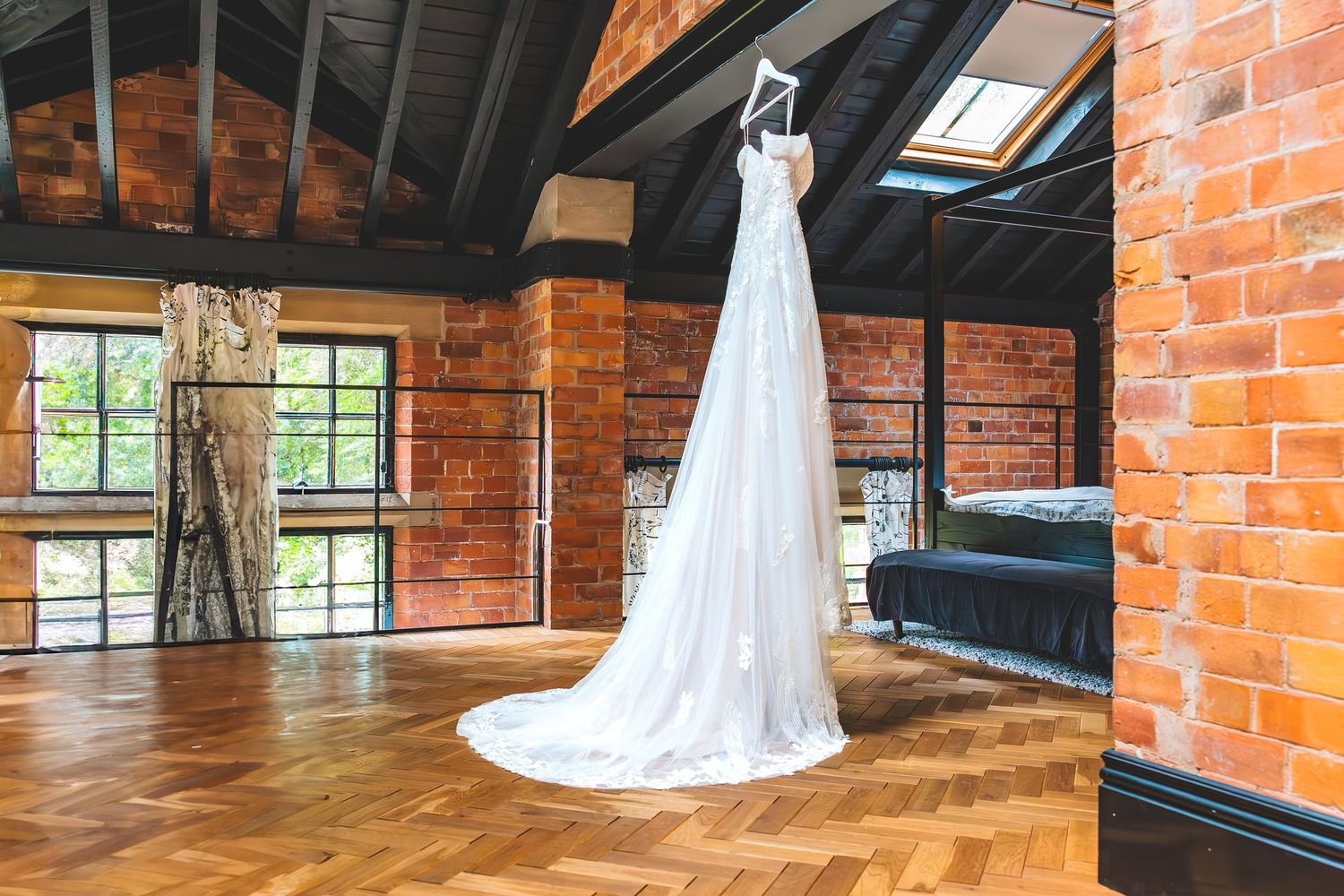 A white wedding dress hangs elegantly in a rustic brick loft space with exposed beams and natural lighting.