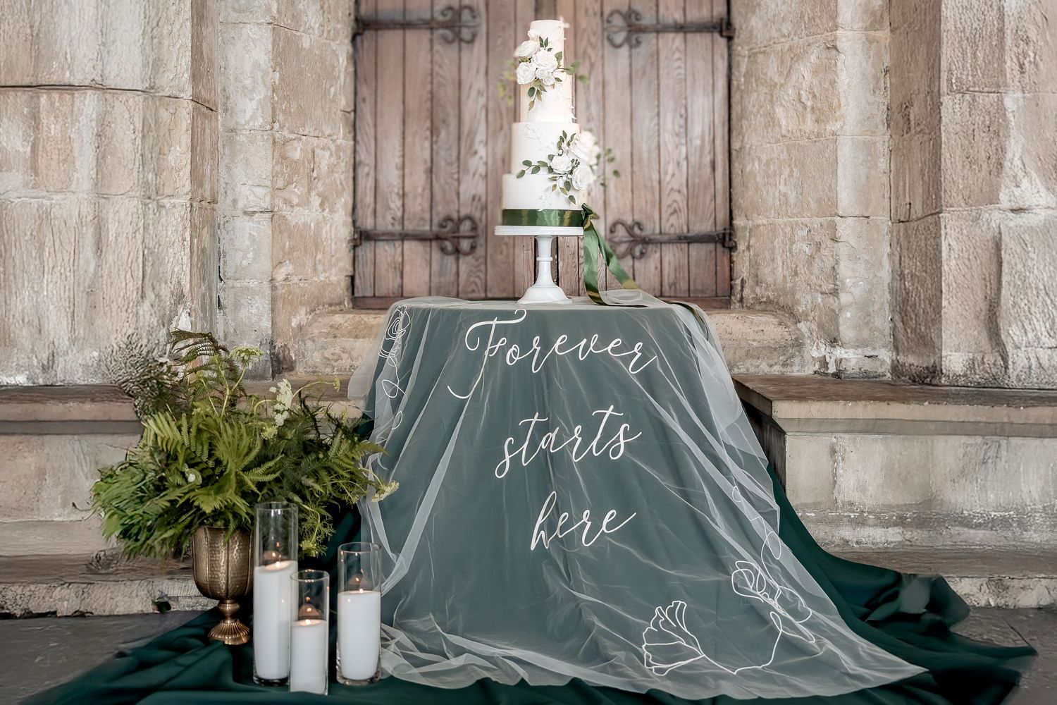 Romantic wedding display with green draping cloth, calligraphy sign reading 'forever starts here', and elegant  flowers.
