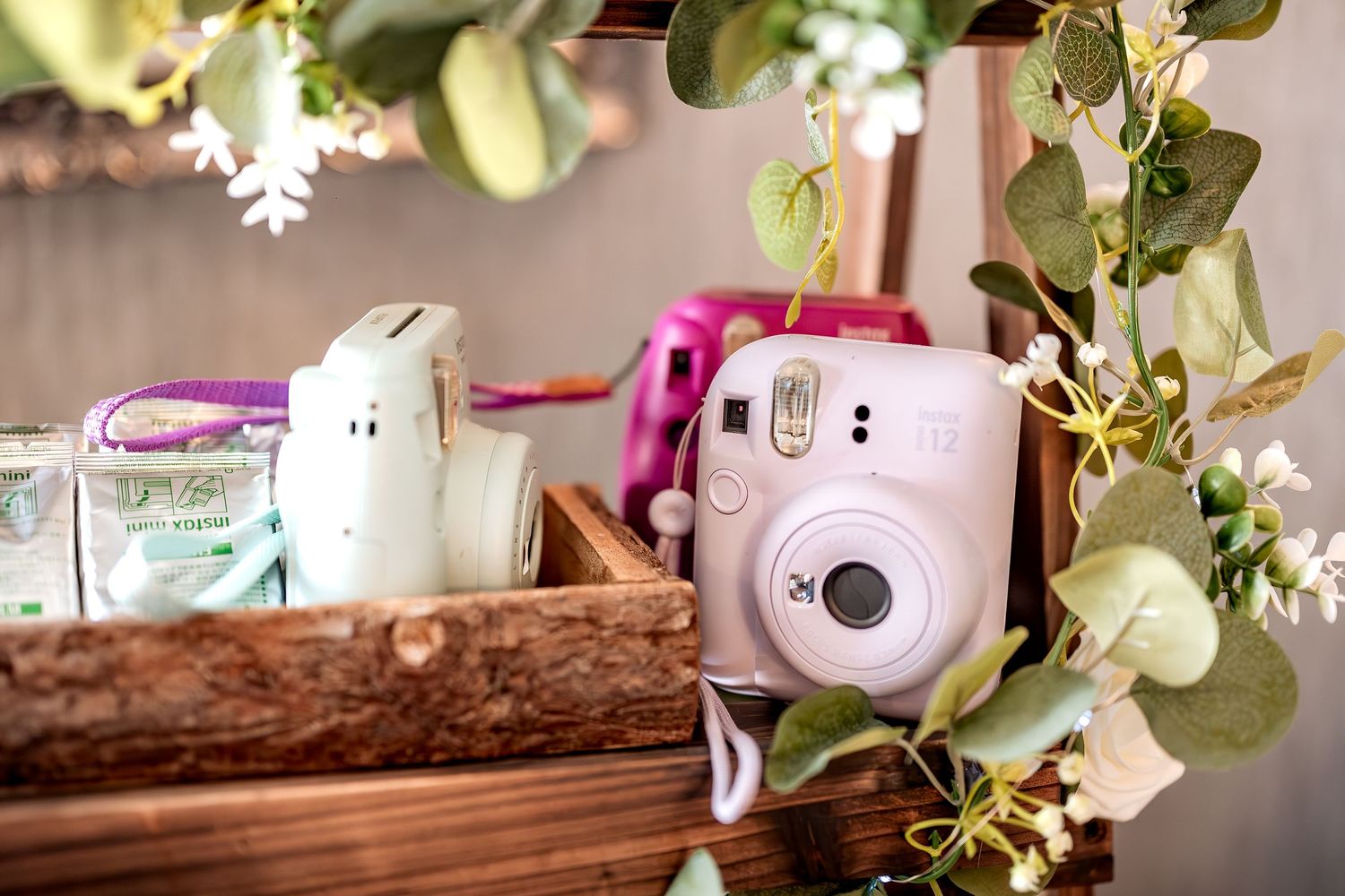 A pink Fujifilm Instax Mini instant camera sits in a wooden box with decorative greenery and white flowers.