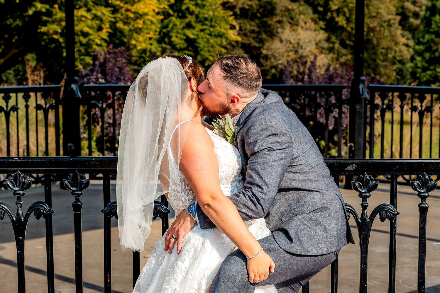 Couple shares romantic wedding day kisses in outdoor garden setting with ornate black fence and trees in background.