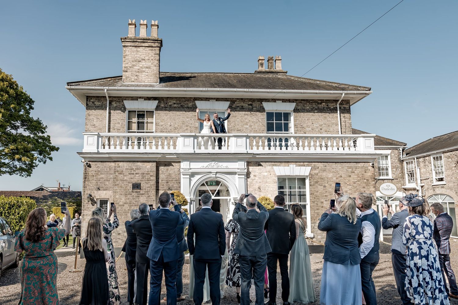 Married couple wave to their guests on their wedding day from a balcony of their wedding venue.