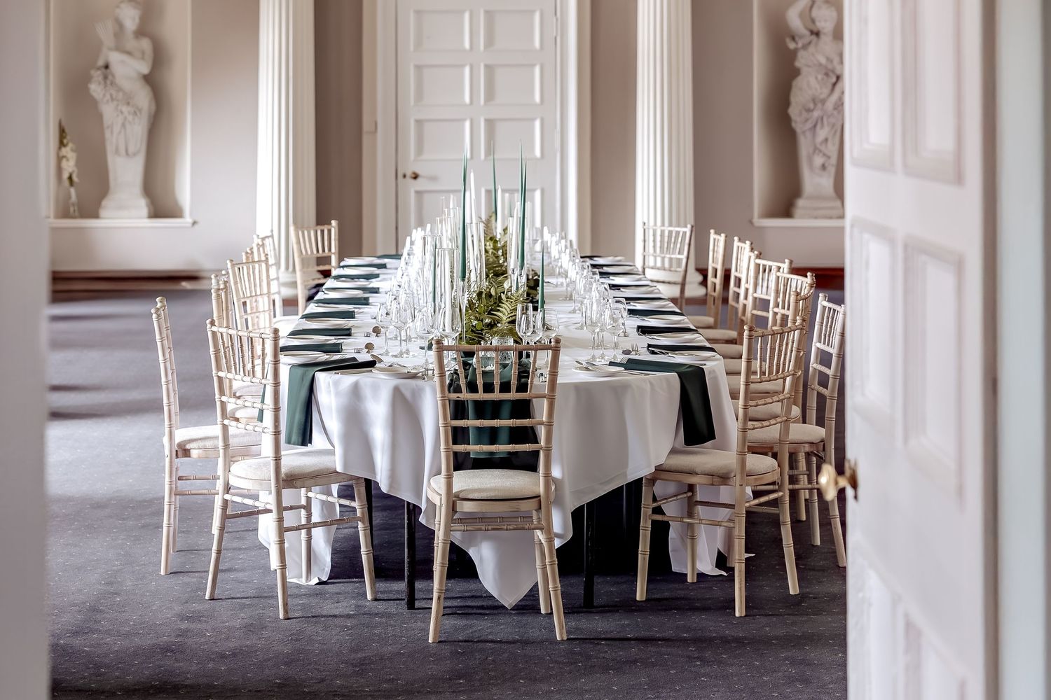 Elegant wedding reception table with white linens, rustic chairs, and classical statues in a formal dining room.