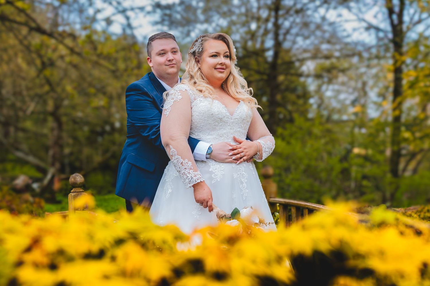 A newly wed couple embraces outdoors surrounded by blooming yellow forsythia bushes and spring trees in a natural setting.